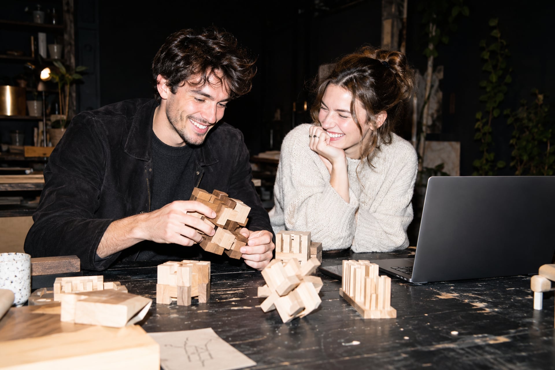Two makers compare wooden assemblies in a Paris atelier after dark.